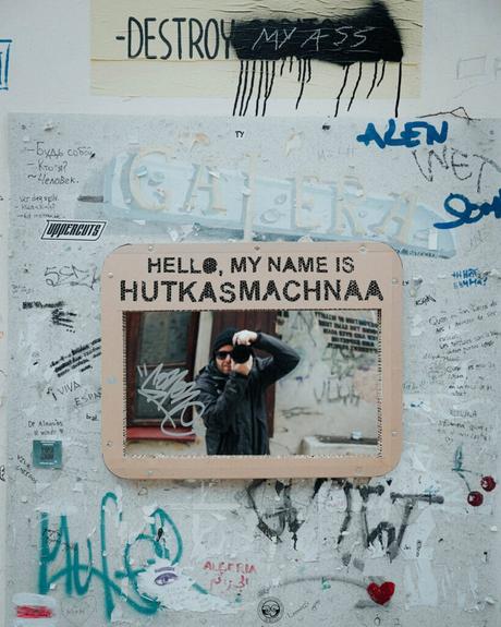 Eine Person mit einer Kamera wird von einem Schild mit der Aufschrift „Hallo, mein Name ist HUTKASMACHNAA“ auf einer Graffitiwand eingerahmt.