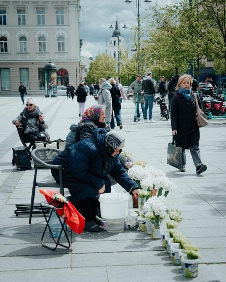 Zwei Frauen verkaufen an einem bewölkten Tag auf einer Stadtstraße Blumen, während Menschen vorbeigehen.