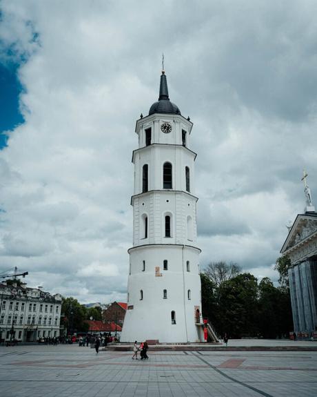 Hoher weißer Uhrenturm mit einer Turmspitze auf einem großen offenen Platz unter einem bewölkten Himmel, Menschen gehen in der Nähe spazieren.