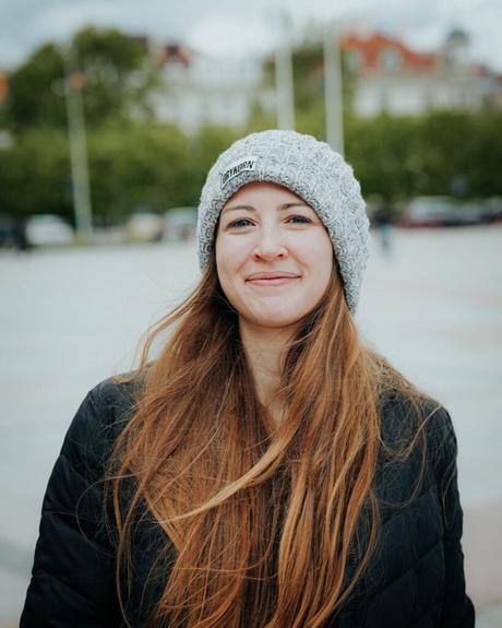 Frau mit langen Haaren, grauer Strickmütze und schwarzer Jacke, lächelt an einem bewölkten Tag im Freien.