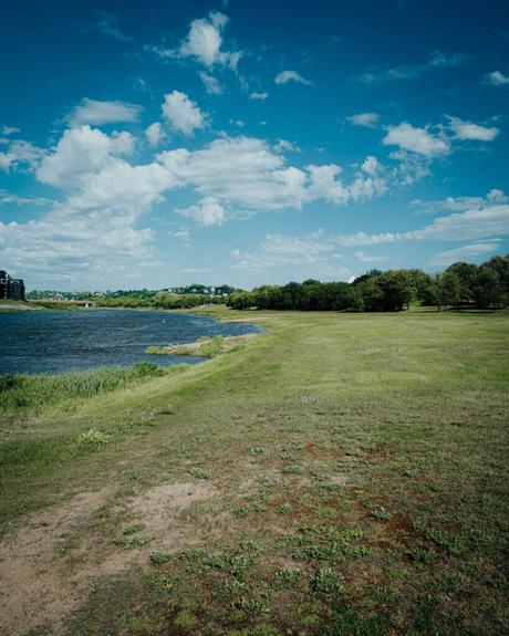 Unter einem strahlend blauen Himmel mit vereinzelten Wolken grenzt eine Grasfläche an einen kleinen See.