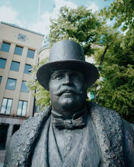 Bronzestatue eines Mannes mit Schnurrbart und Zylinder vor einem Gebäude und grünen Bäumen.