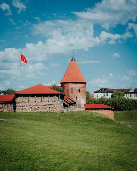 Ein Backsteinschloss mit rotem Dach, Turm und Flagge auf einem grasbewachsenen Hügel unter einem blauen Himmel mit Wolken.