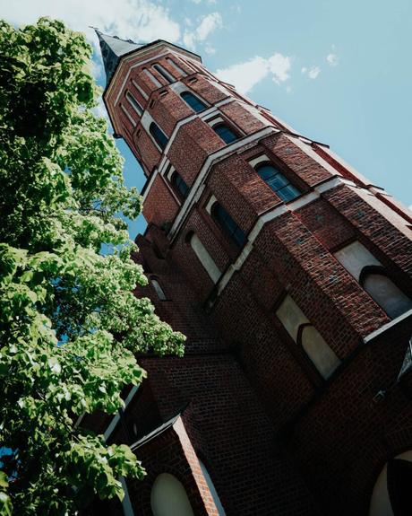 Ein hoher Kirchturm aus rotem Backstein mit Bogenfenstern neben grünen Bäumen, von unten betrachtet.