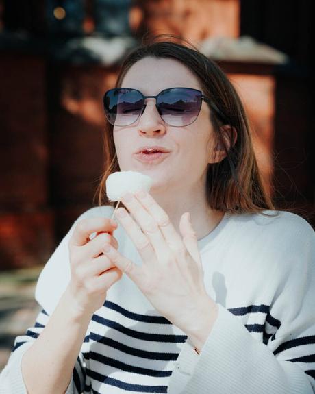 Frau mit Sonnenbrille, lächelnd und mit einem weißen Leckerbissen am Stiel in der Hand, trägt einen gestreiften Pullover im Freien.