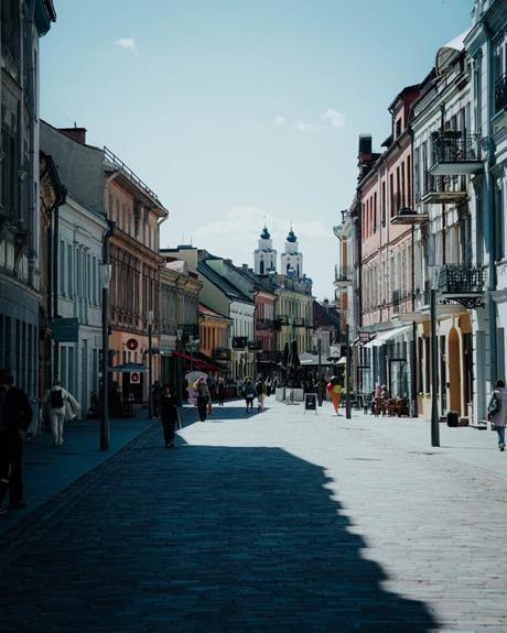Eine sonnenbeschienene europäische Straße mit farbenfrohen Gebäuden und Passanten, im Hintergrund sind Kirchtürme zu sehen.