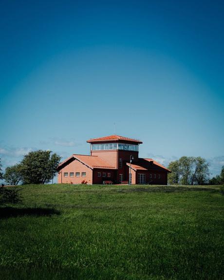 Ein rotes Backsteinhaus mit Turm steht auf einer Wiese unter einem klaren blauen Himmel.