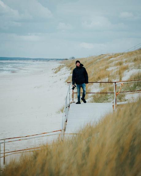 Eine Person in einer dunklen Jacke geht unter einem bewölkten Himmel allein auf einem Holzsteg an einem sandigen, grasbewachsenen Strand entlang.