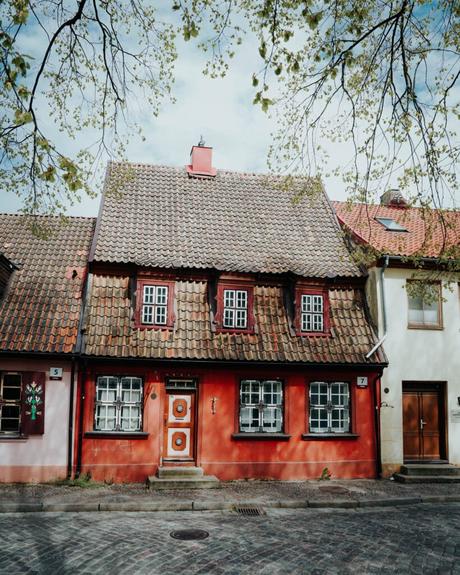 Ein malerisches rotes Haus mit Ziegeldach und Dachgauben in einer Kopfsteinpflasterstraße unter belaubten Ästen.