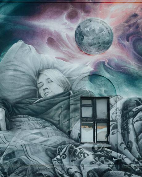 Ein Wandgemälde einer schlafenden Frau mit einem Kissen, einer Decke und einem Mond darüber, gemalt um ein echtes Fenster.