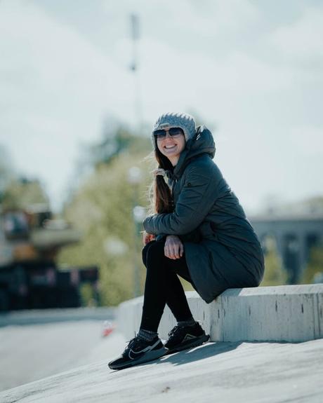 Eine lächelnde Frau in Mantel und Mütze sitzt an einem sonnigen Tag im Freien auf einem Betonvorsprung.