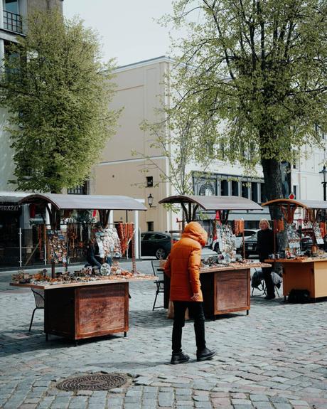 Eine Person in einem orangefarbenen Mantel geht an Marktständen im Freien auf einer Kopfsteinpflasterstraße mit Bäumen und Gebäuden vorbei.