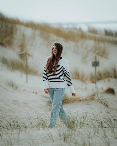 Frau mit langen Haaren und gestreiftem Pullover läuft an einem bewölkten Tag über Sanddünen mit Gras.