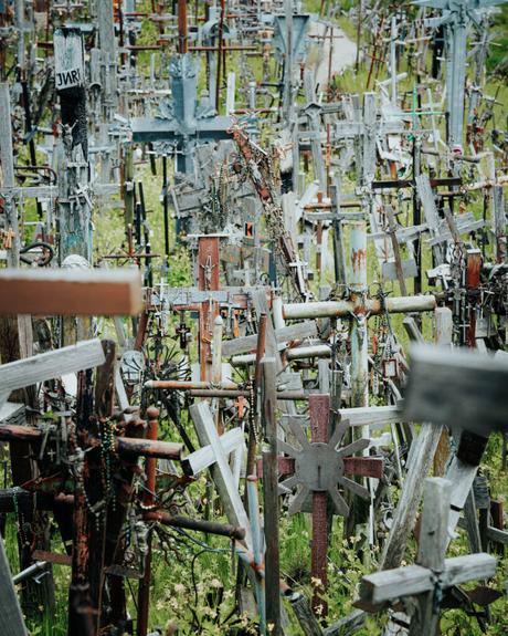 Dichte Ansammlung verwitterter Holzkreuze, einige mit Rosenkränzen, in einer überwucherten Grasfläche.
