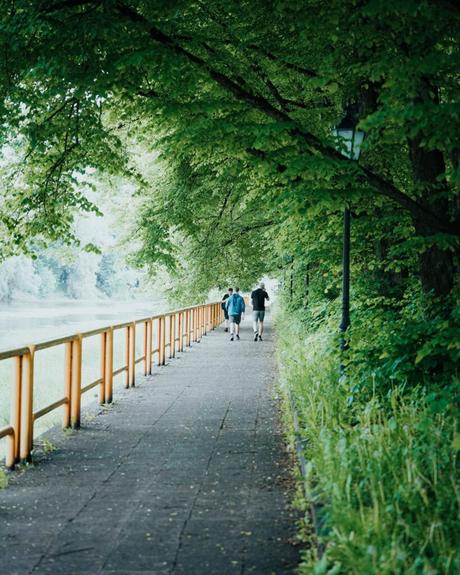 Zwei Menschen gehen einen von Bäumen gesäumten Weg neben einem Fluss entlang, der von einem gelben Geländer begrenzt wird.