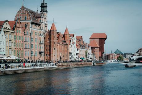 An einem ruhigen, sonnigen Tag säumen farbenfrohe historische Gebäude und ein Backsteinturm eine Uferpromenade.