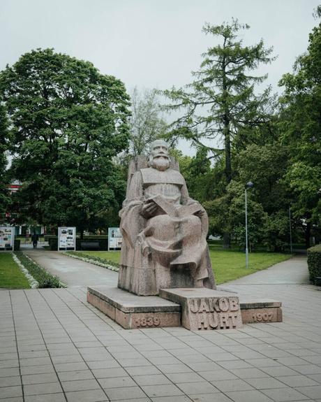 Eine sitzende Steinstatue von Jakob Hurt in einem von Bäumen und Wegen umgebenen Park.