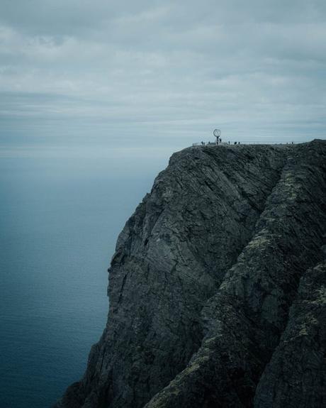Menschen stehen in der Nähe einer Weltkugel-Skulptur auf einer dramatischen Klippe über dem Meer unter einem bewölkten Himmel.