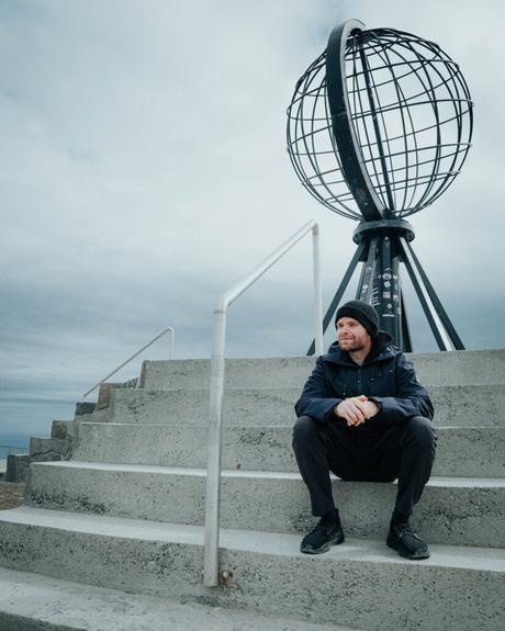 Ein Mann sitzt auf Betonstufen unter einer großen Metallkugel-Skulptur unter einem bewölkten Himmel.