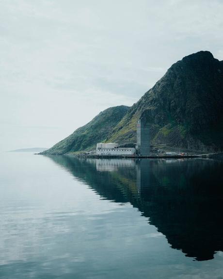 Ein Gebäude und ein Silo liegen am Fuße eines steilen, felsigen Berges, in dem sich das Wasser klar spiegelt.