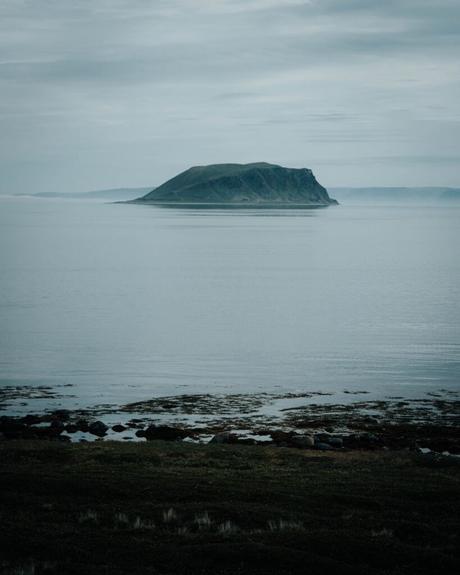 Eine einsame grüne Insel liegt in ruhigem, nebligem Wasser unter einem bedeckten Himmel, gesehen von einem felsigen Ufer aus.
