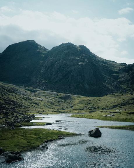 Ein Fluss schlängelt sich durch ein grünes Tal mit felsigen Bergen im Hintergrund unter einem bewölkten Himmel.