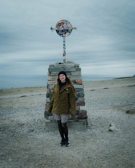 Eine Frau steht lächelnd vor einem verzierten steinernen Wegweiser an einem felsigen, bewölkten Strand.