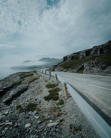 Eine kurvenreiche Küstenstraße mit Leitplanke, felsigen Klippen und nebligen Bergen unter einem bewölkten Himmel.