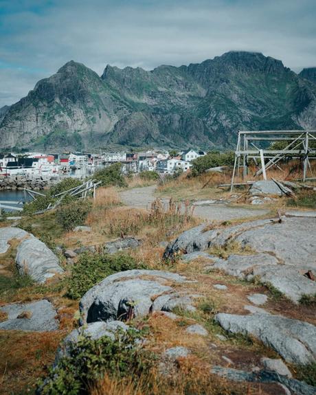 Felsiger Weg mit Gras, entfernte Häuser und große Berge unter einem bewölkten Himmel in einem Küstendorf.
