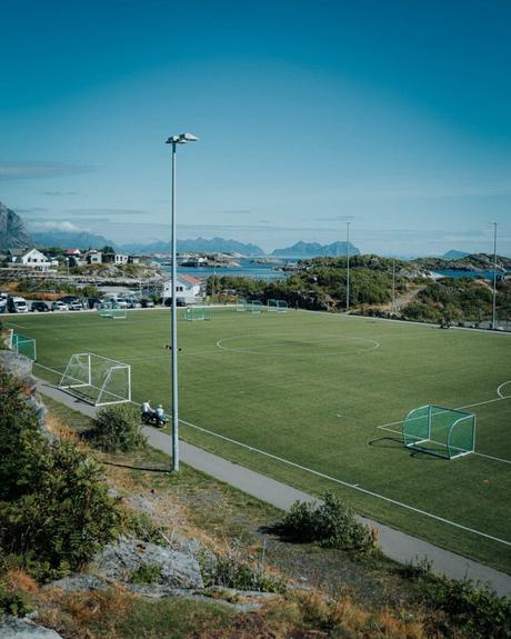Fußballplatz mit grünen Toren, umgeben von Natur, Häusern und Bergen unter einem klaren blauen Himmel.