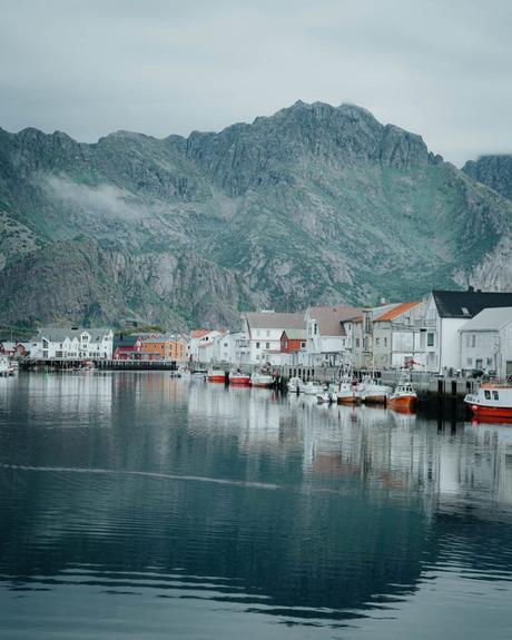 Bunte Häuser und Boote säumen eine ruhige Uferpromenade mit Bergen im Hintergrund unter einem bewölkten Himmel.