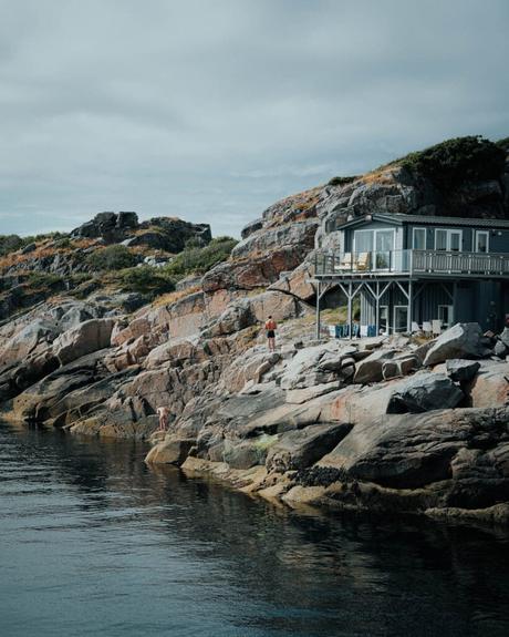 Haus am felsigen Ufer mit Menschen, die auf Felsen klettern, und ruhigem Wasser im Vordergrund unter bewölktem Himmel.