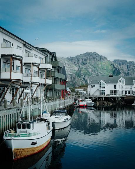 An einem ruhigen Fjord mit Bergen im Hintergrund und blauem Himmel liegen Boote an Holzhäusern an.