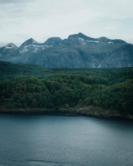 Bergkette mit Schneeflecken, dichter grüner Wald und ruhiges Wasser im Vordergrund unter einem bewölkten Himmel.