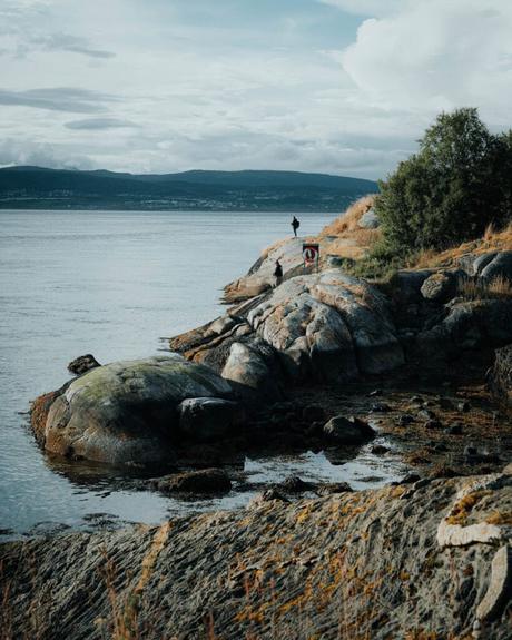 Eine Person steht an einem felsigen Ufer am ruhigen Wasser, mit Bergen und Wolken im Hintergrund.