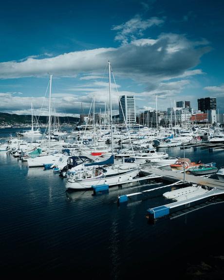 Boote und Yachten in einem Jachthafen mit städtischen Gebäuden und Bergen im Hintergrund unter einem bewölkten Himmel.