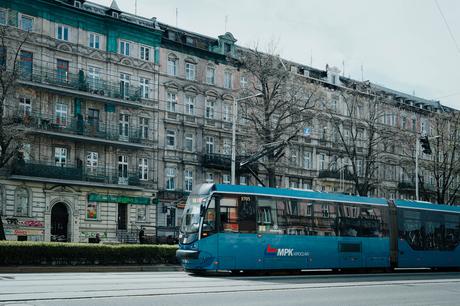 Blaue Straßenbahn, die an alten Wohnhäusern mit Balkonen und Bäumen auf einer Stadtstraße vorbeifährt.