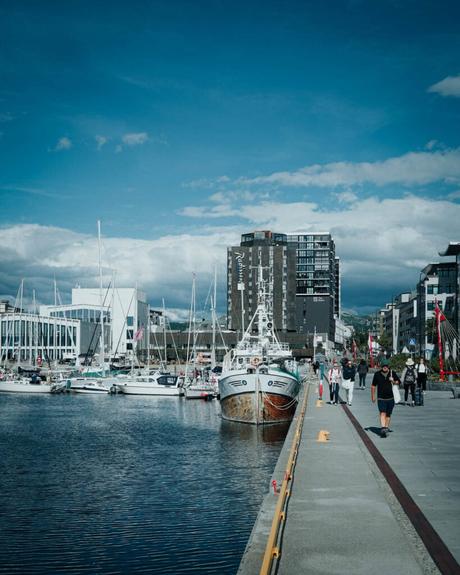 Angedockte Boote an einer Uferpromenade mit Spaziergängern in der Nähe und modernen Gebäuden im Hintergrund.