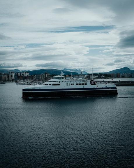 Eine große Fähre fährt über ein Gewässer mit einer Stadtlandschaft und Bergen im Hintergrund unter einem bewölkten Himmel.