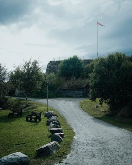 Ein Kiesweg führt zu einem steinernen Gebäude mit einer Fahne; Bänke und Bäume säumen die Rasenfläche.