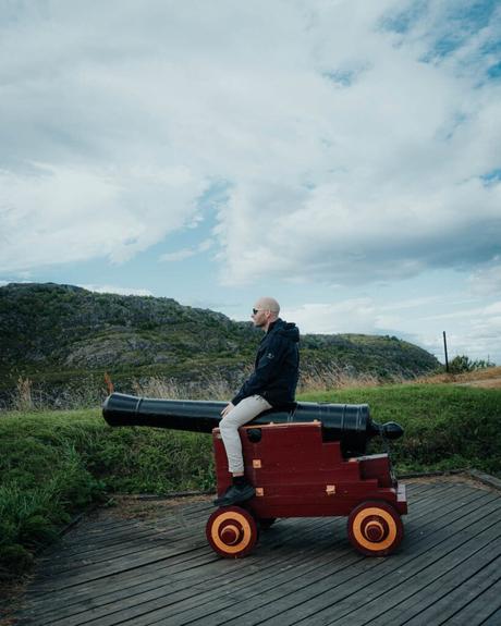 Ein Mann sitzt auf einer roten, dekorativen Kanone im Freien mit Hügeln und bewölktem Himmel im Hintergrund.