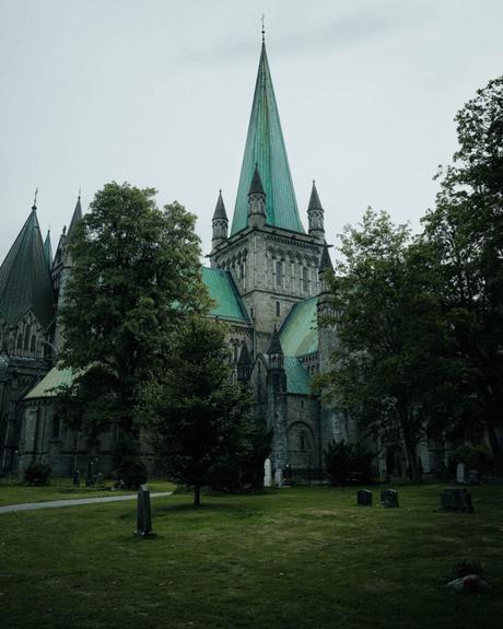 Eine große Steinkathedrale mit einem grünen Turm, umgeben von Bäumen und Grabsteinen an einem bewölkten Tag.