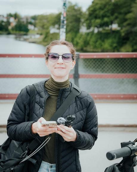 Eine Frau mit Sonnenbrille und schwarzer Jacke steht auf einer Brücke, lächelt und hält ein Telefon.