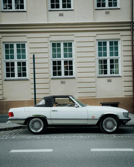 Ein klassisches weißes Cabrio, das vor einem beigen Gebäude mit großen Fenstern geparkt ist.