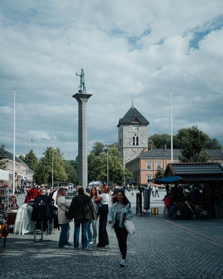 Menschen beim Spazierengehen und Einkaufen auf einem Stadtplatz mit einer großen Statue und einem Uhrenturm im Hintergrund.
