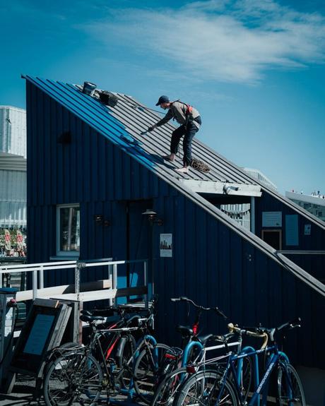 Eine Person mit einer Mütze arbeitet auf dem schrägen Dach eines blauen Gebäudes, unter dem Fahrräder geparkt sind.