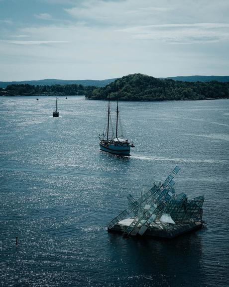 Ein Segelboot auf einer glitzernden blauen Bucht passiert eine moderne Glasskulptur mit einer grünen Insel im Hintergrund.