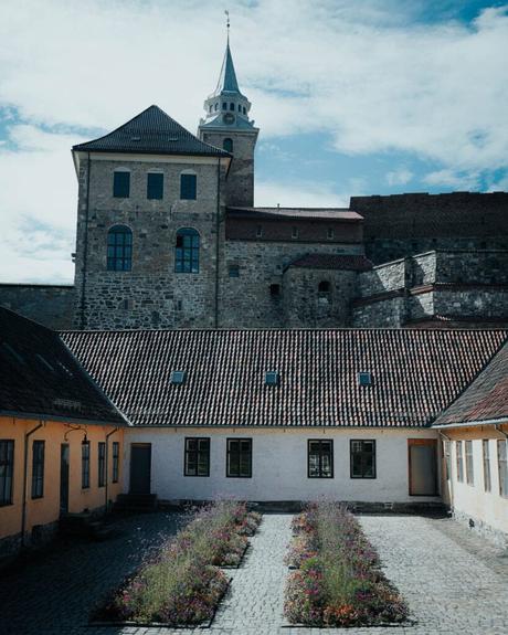 Ein steinernes Schloss mit einem hohen Turm hinter einem Hofgarten mit gepflasterten Wegen und Blumenbeeten.