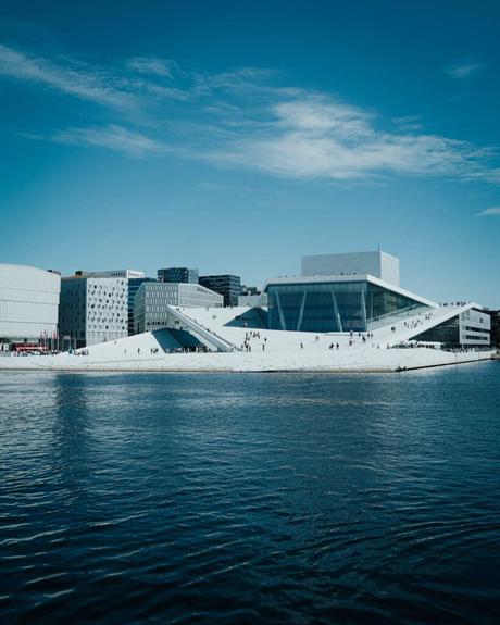 Modernes weißes Gebäude mit schrägem Dach am Wasser unter einem klaren blauen Himmel, Menschen gehen auf dem Dach.