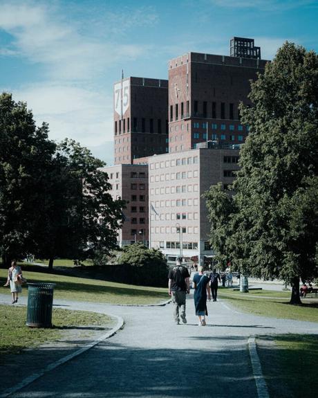 Zwei Personen gehen auf einem Parkweg auf ein großes Backsteingebäude mit der Nummer 75 zu, das von Bäumen umgeben ist.
